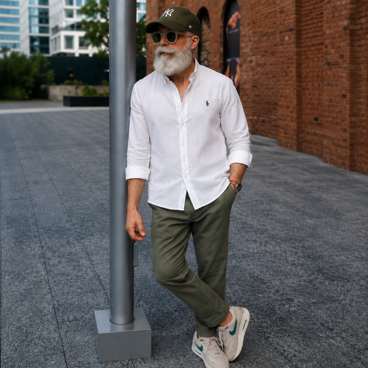Older man with a white beard wearing a white shirt and olive trousers leaning against a pole.