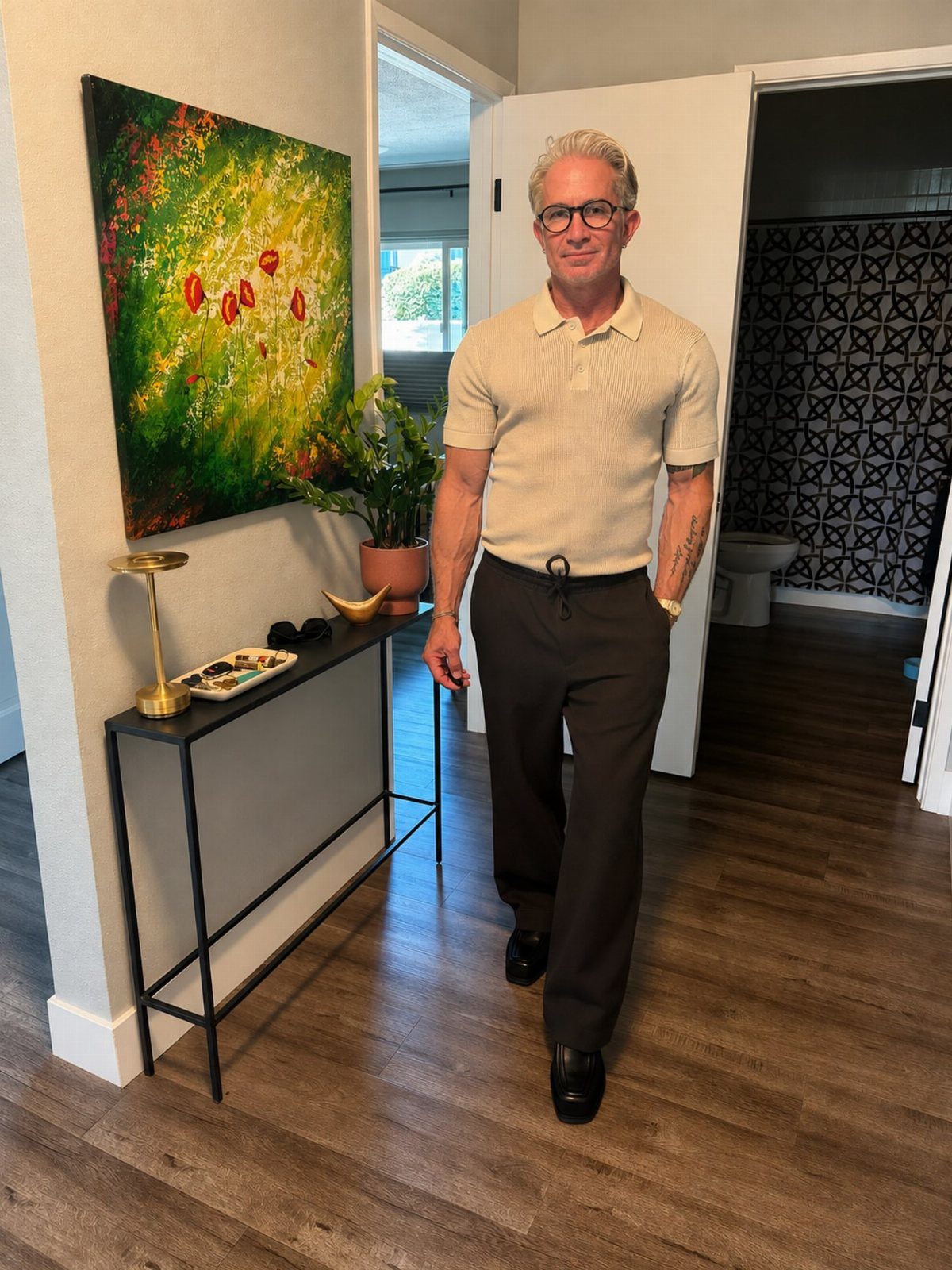 A smiling middle-aged man wearing a beige polo shirt and dark trousers standing in a hallway.