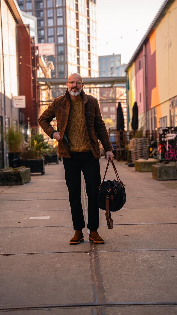 A mature man wearing a brown jacket, yellow sweater, black pants, and brown boots standing outdoors.