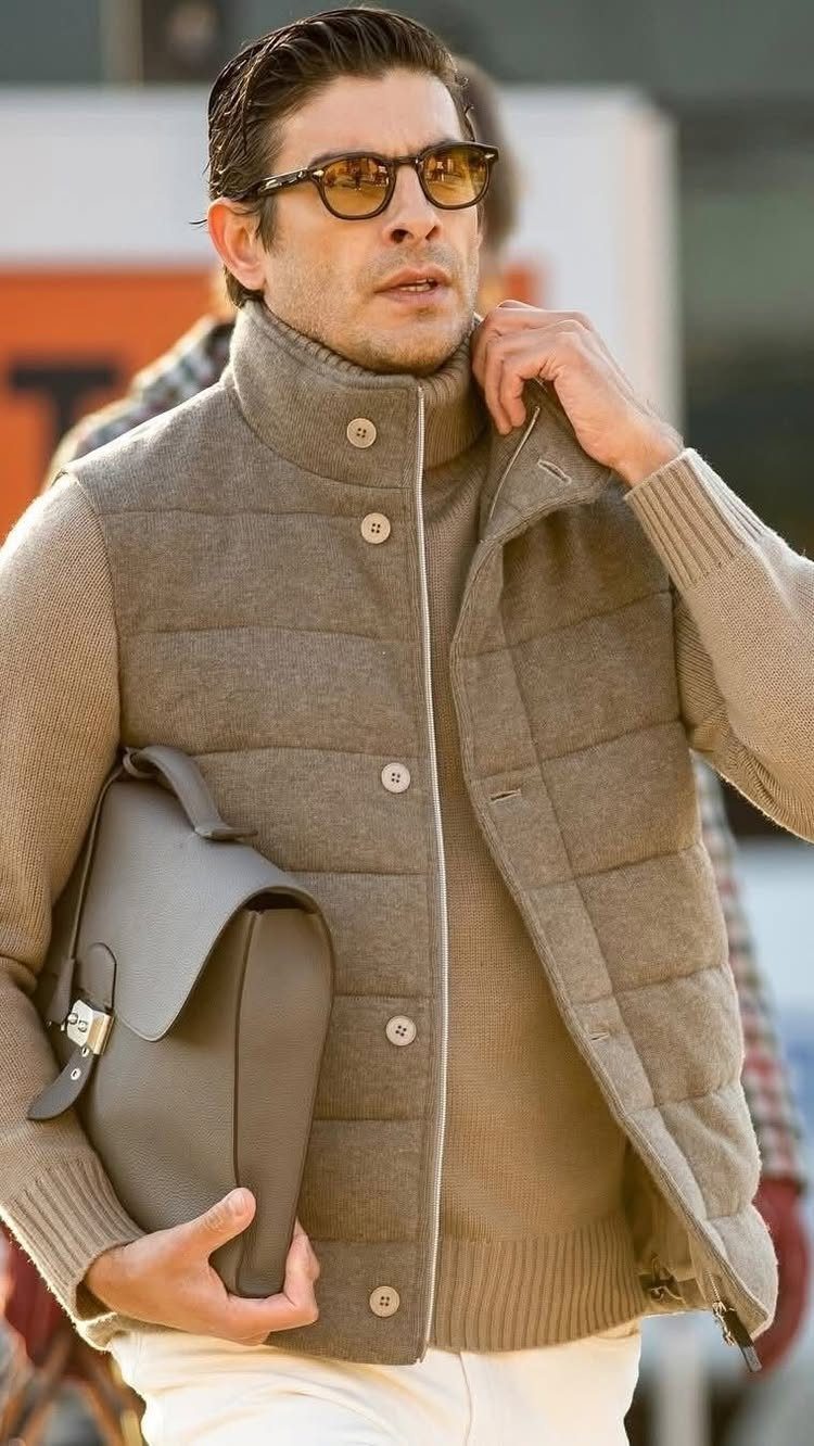 Man in a tan turtleneck and quilted vest carrying a grey leather briefcase outdoors.
