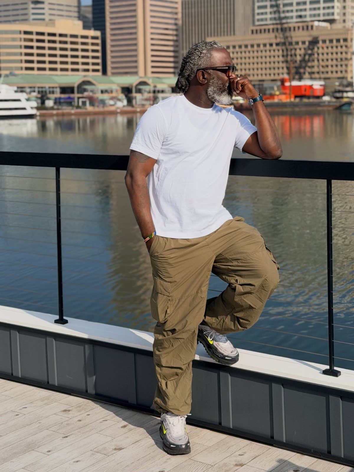 A mature man in a white t-shirt and khaki cargo pants posing by a city waterfront.