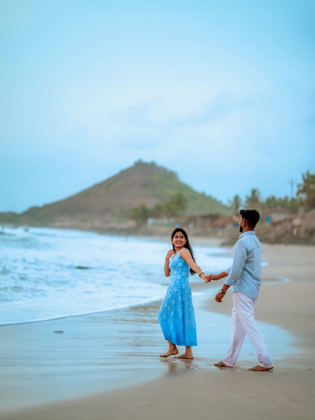 A man in a light blue shirt and white pants holds hands with a woman in a blue dress on a beach.