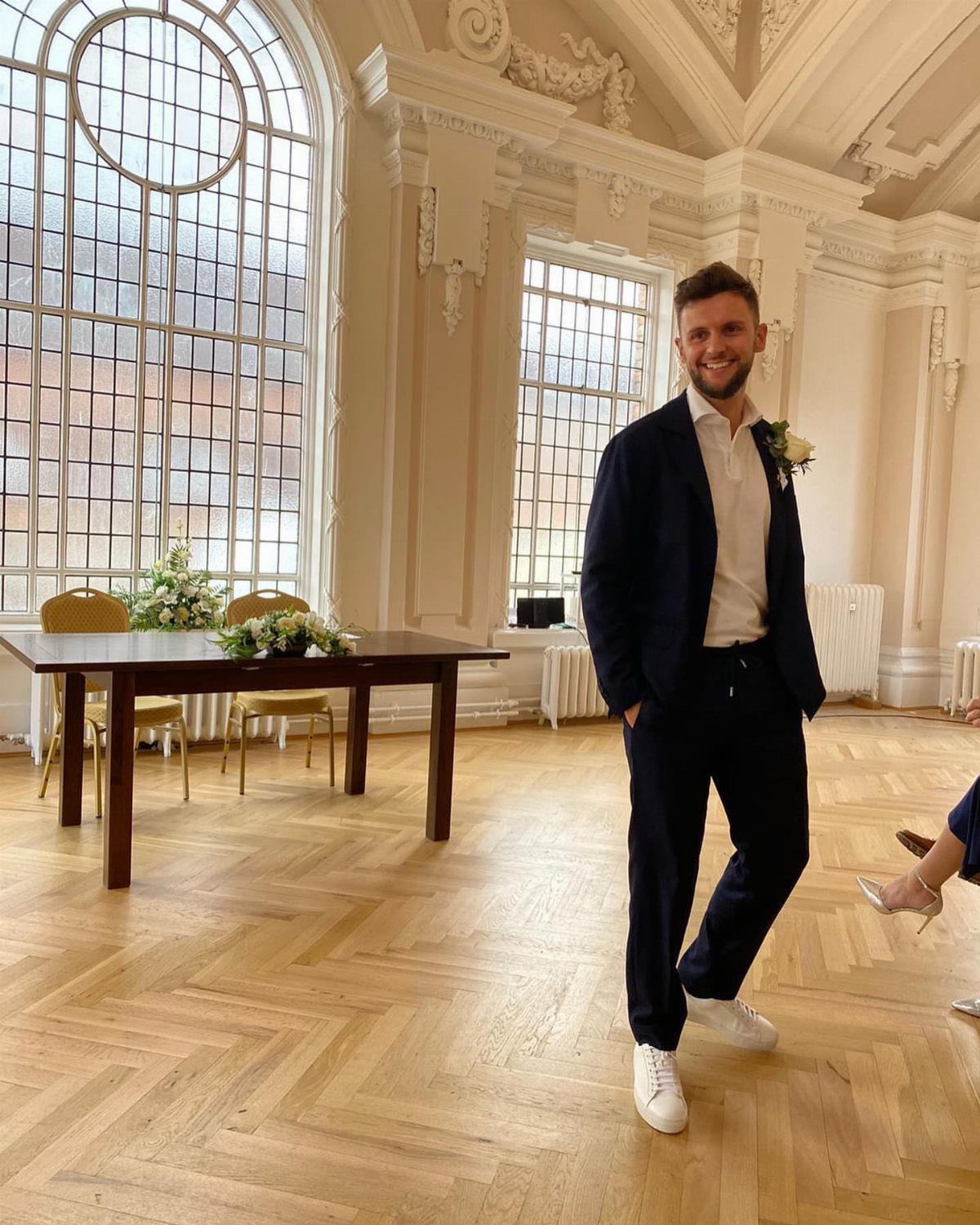 A smiling man wearing a navy blazer, white shirt, and dark navy trousers in a bright room.