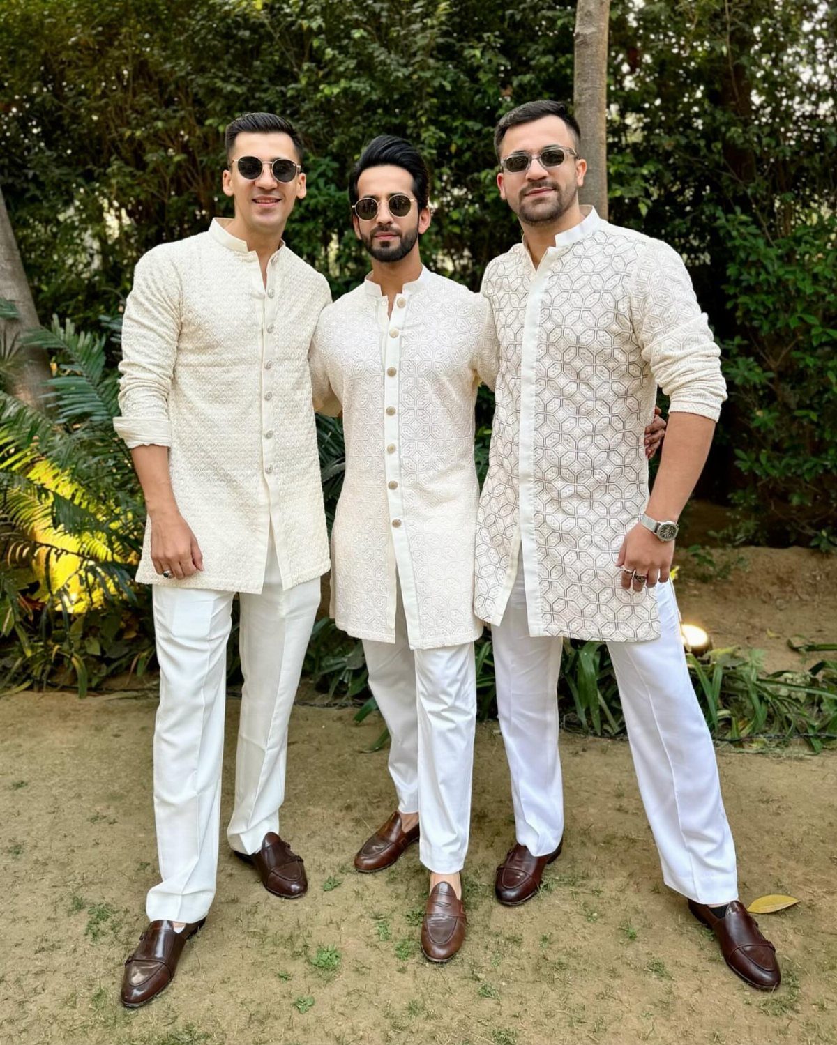 Three men in cream patterned tunics and white trousers wearing dark brown leather loafers outdoors.