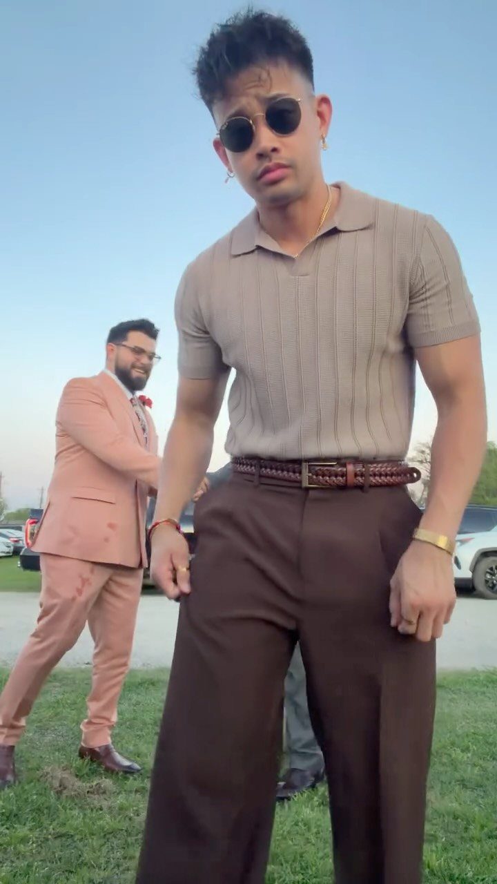 Man in a tan polo and brown trousers posing outdoors with another man in a pink suit.
