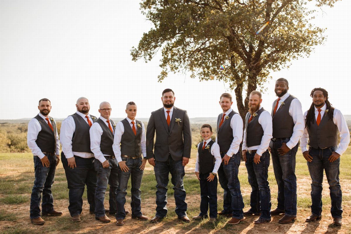Group of men and a boy wearing vests, ties, jeans, and boots standing outdoors.