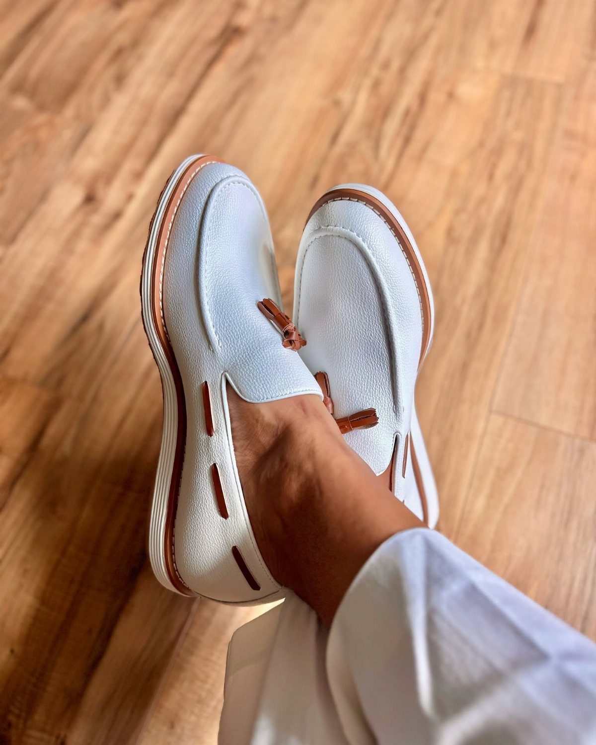 White slip-on loafers with brown trim worn on a person's feet on wooden flooring.