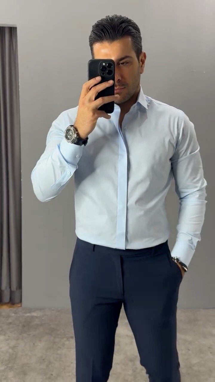 Man in a light blue button-down shirt and navy trousers taking a mirror selfie.