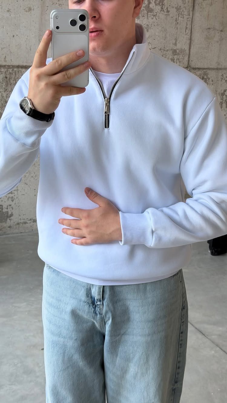 A man in a white quarter-zip sweatshirt and light blue jeans taking a mirror selfie.