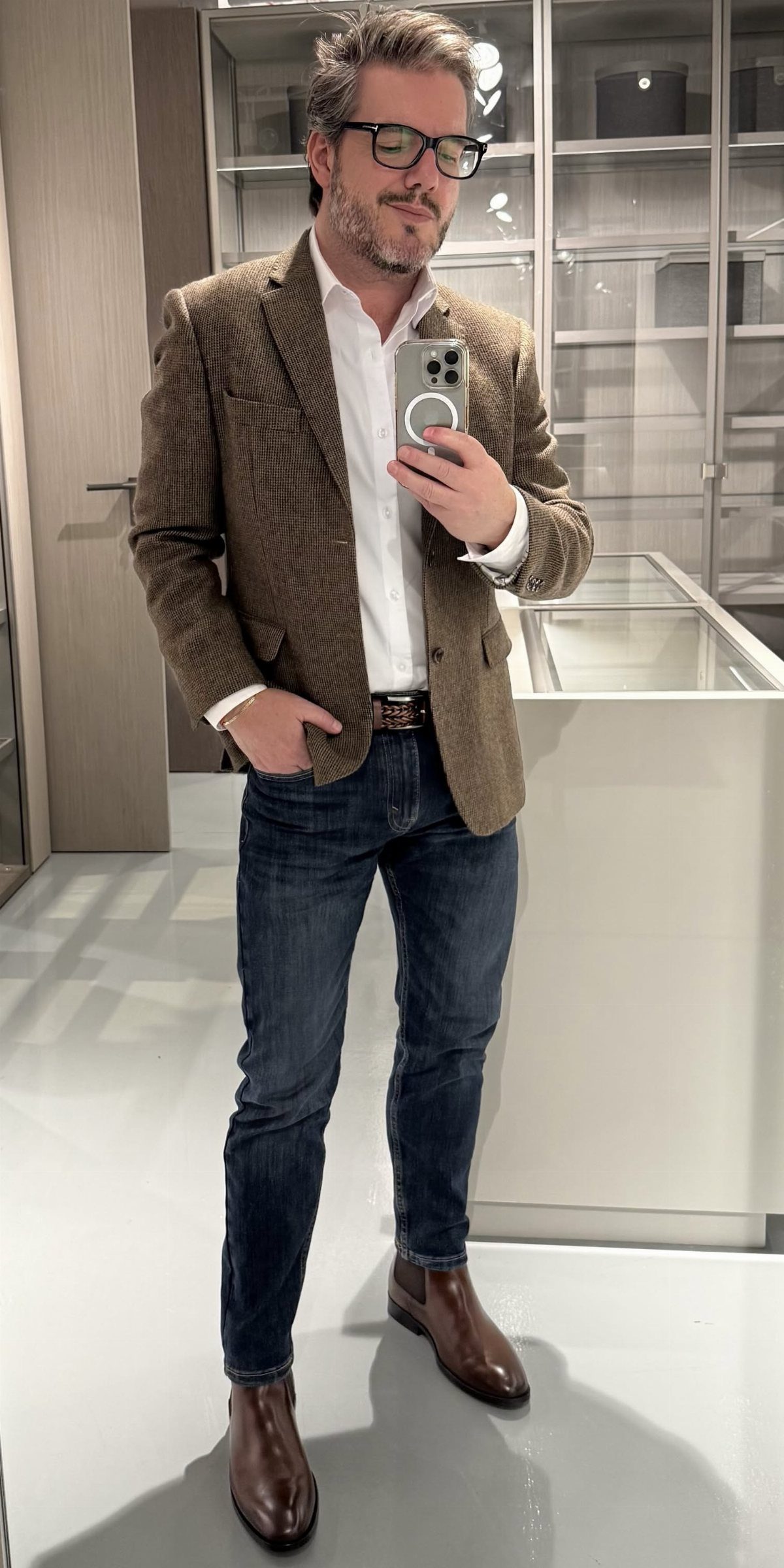 Man in a brown blazer, white shirt, jeans, and brown boots posing for a mirror selfie.