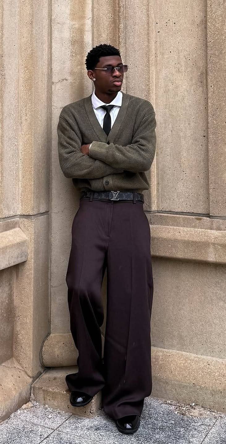 A young man in a brown cardigan, tie, and wide-leg trousers posing against a stone wall.