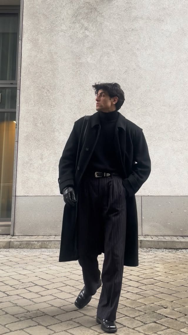 A man wearing a black turtleneck, long coat, and trousers standing against a grey wall.