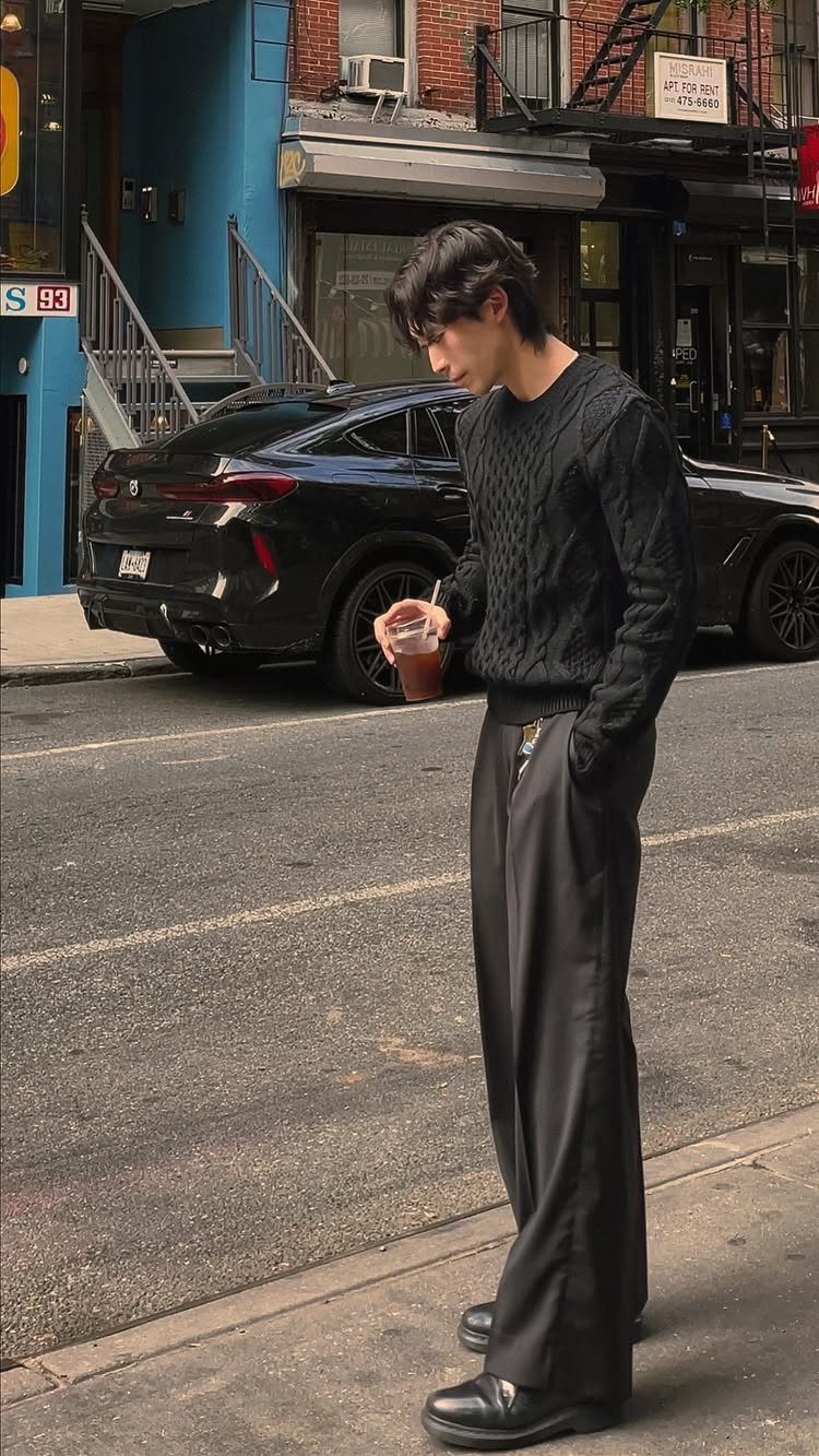 A young man in an all-black monochrome outfit standing on a city street next to a black car.
