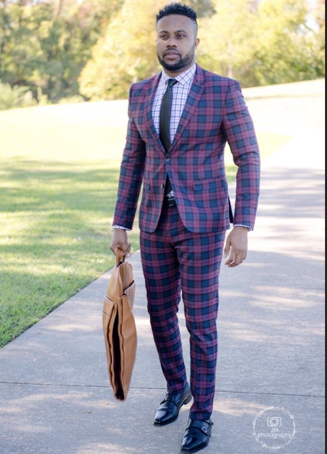 Man in a blue and red plaid suit walking outdoors while carrying a tan leather briefcase.