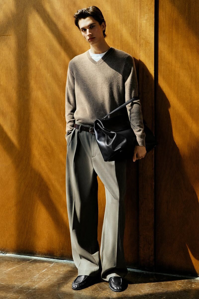 Man wearing a taupe sweater and olive green wide leg pants standing against a wooden wall.
