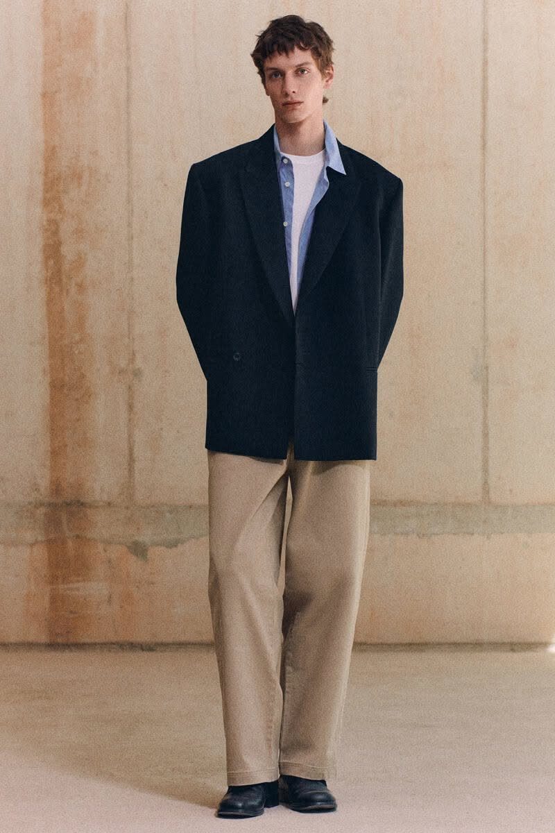 A man wearing a navy blazer, light blue shirt, and tan wide-leg trousers standing against a wall.
