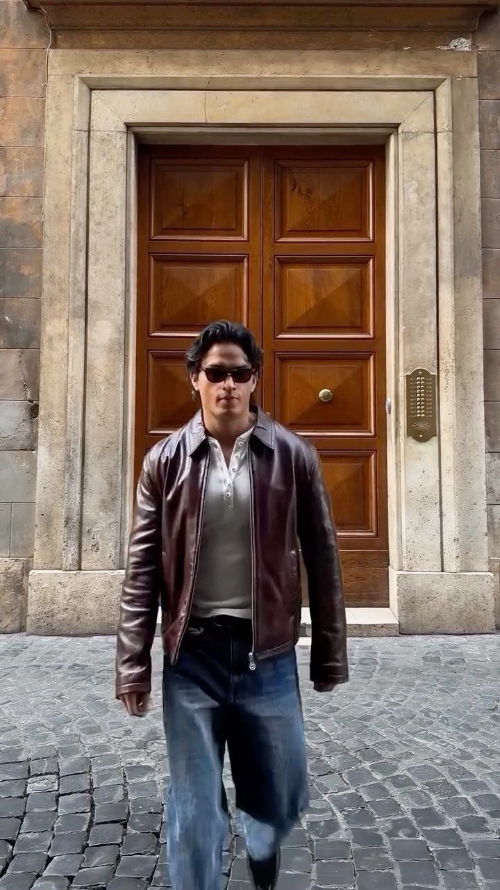 A man walking outdoors wearing a dark brown leather jacket, white shirt, and blue jeans.