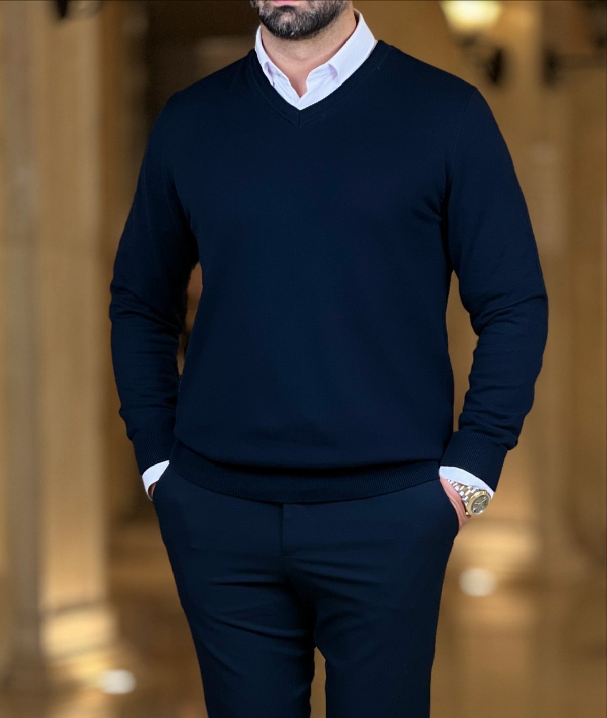 A man wearing a navy blue V-neck sweater over a white collared shirt and navy pants.