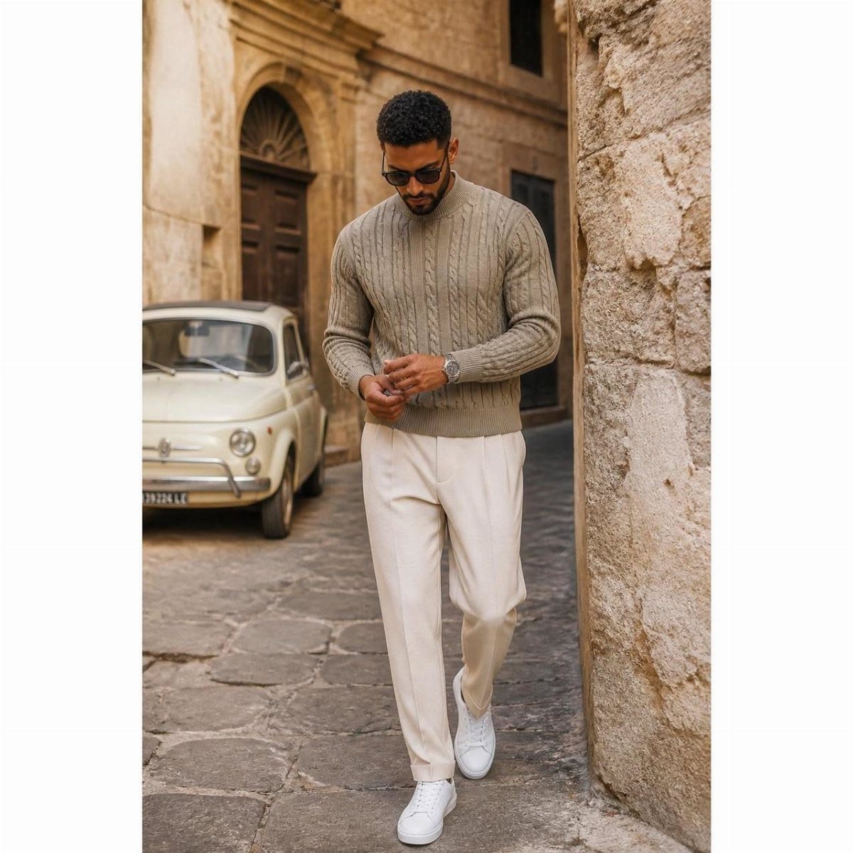 Man walking in a tan cable-knit sweater and cream trousers on a cobblestone street.