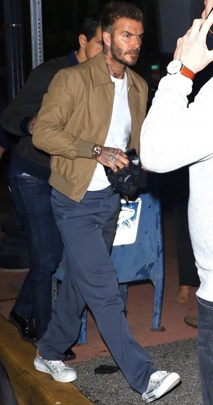 Man walking in a tan jacket, white t-shirt, grey trousers, and white sneakers.