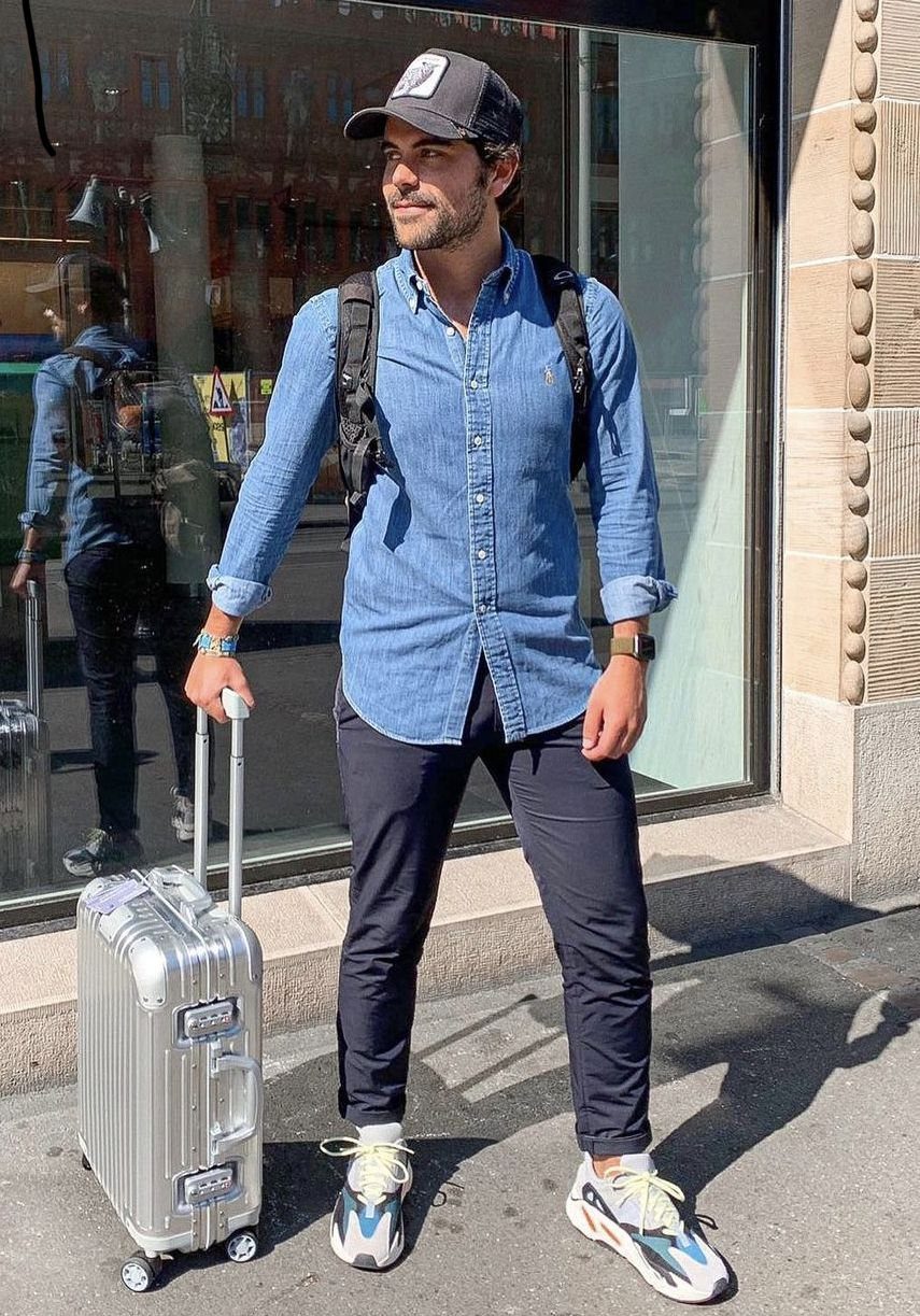 Man in a blue button-down shirt and dark trousers standing with a silver suitcase.