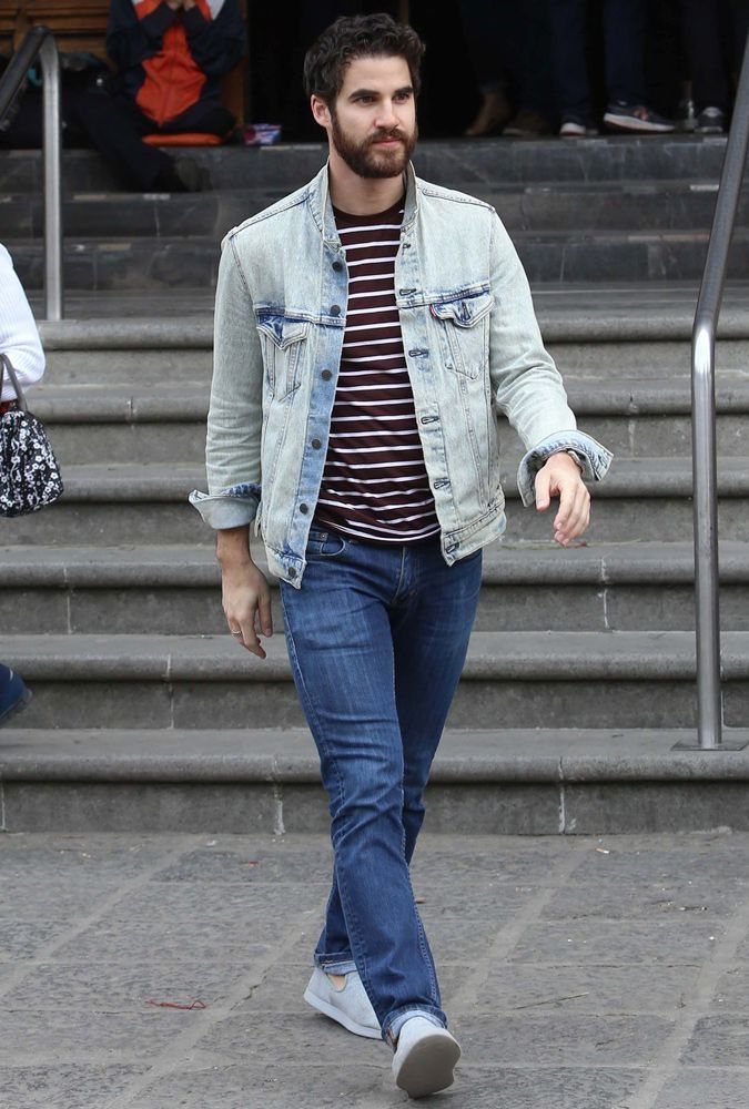 A man walking down stairs wearing a denim jacket, striped shirt, jeans, and slip-on sneakers.