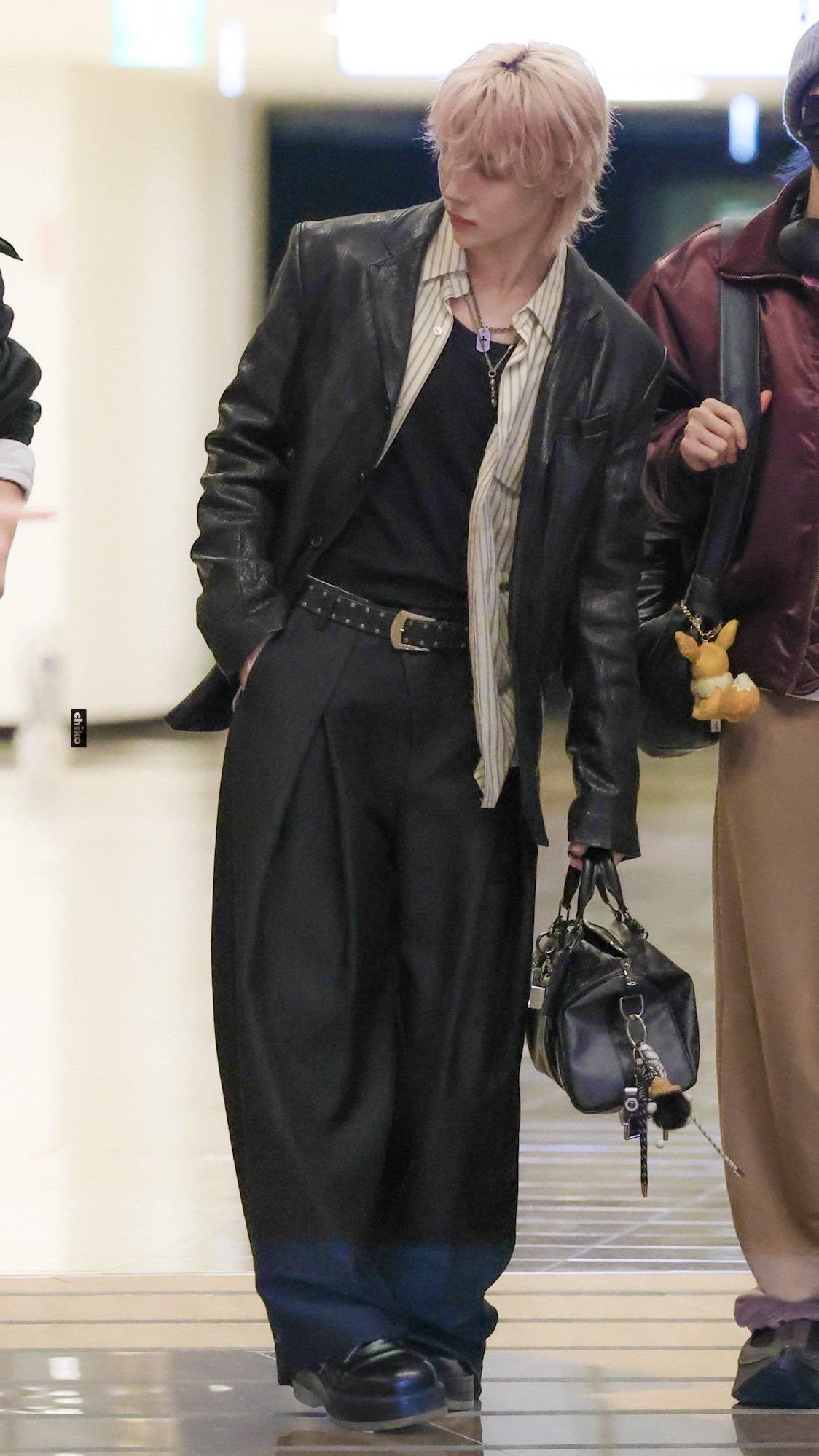 A person in a black leather blazer and wide-leg trousers walking through an airport terminal.