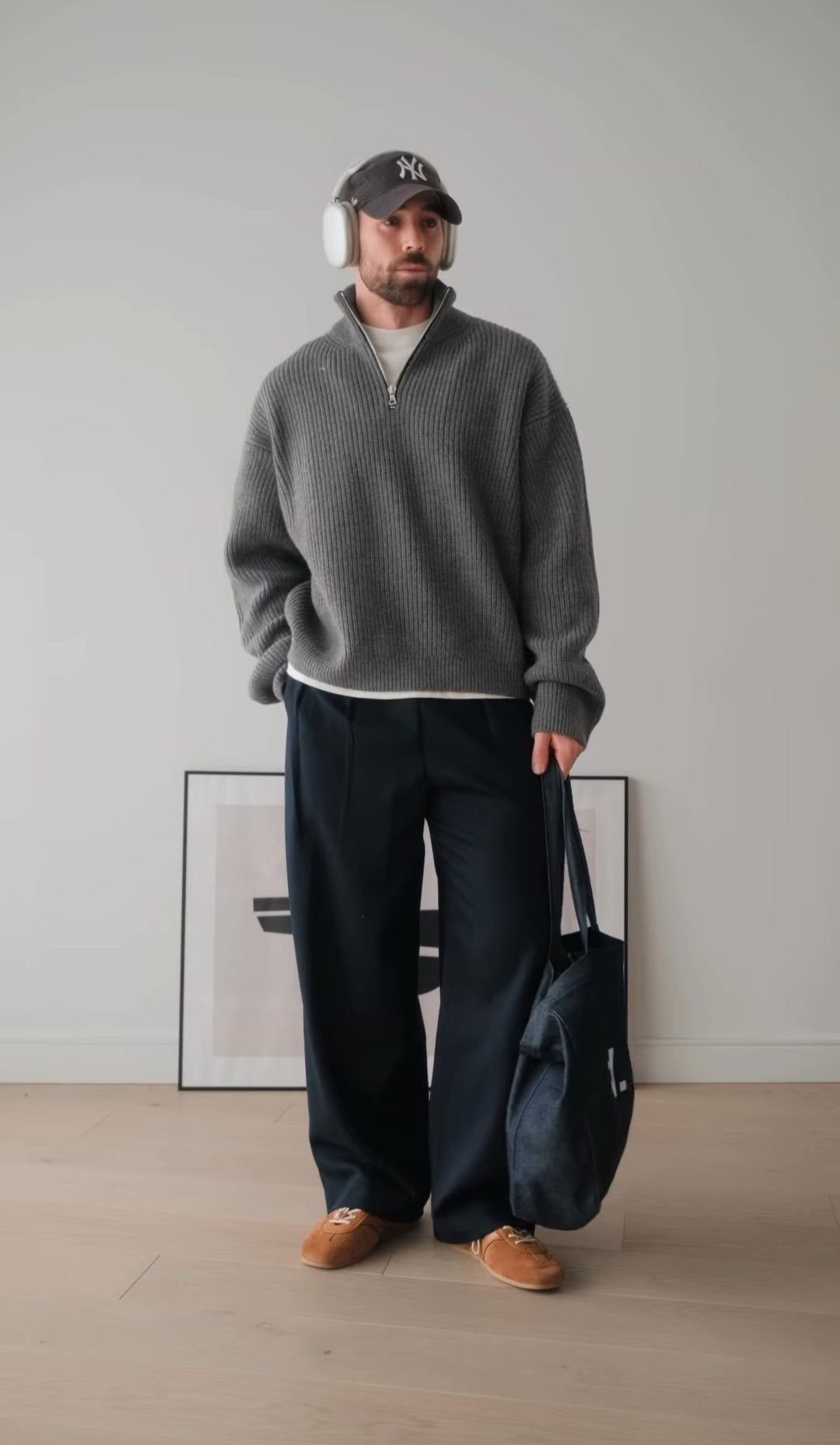A man in a grey sweater, navy trousers, and tan loafers holding a tote bag.