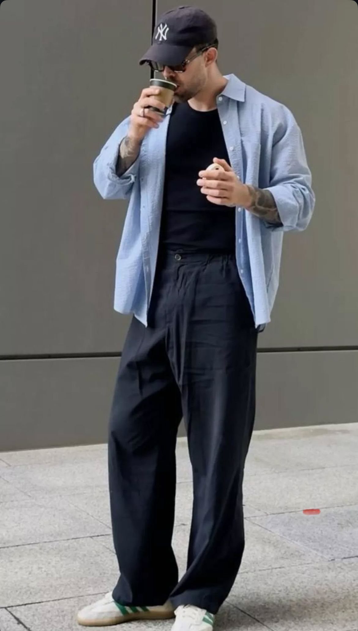 Man wearing black wide-leg trousers, a light blue shirt, and a black baseball cap while holding coffee.