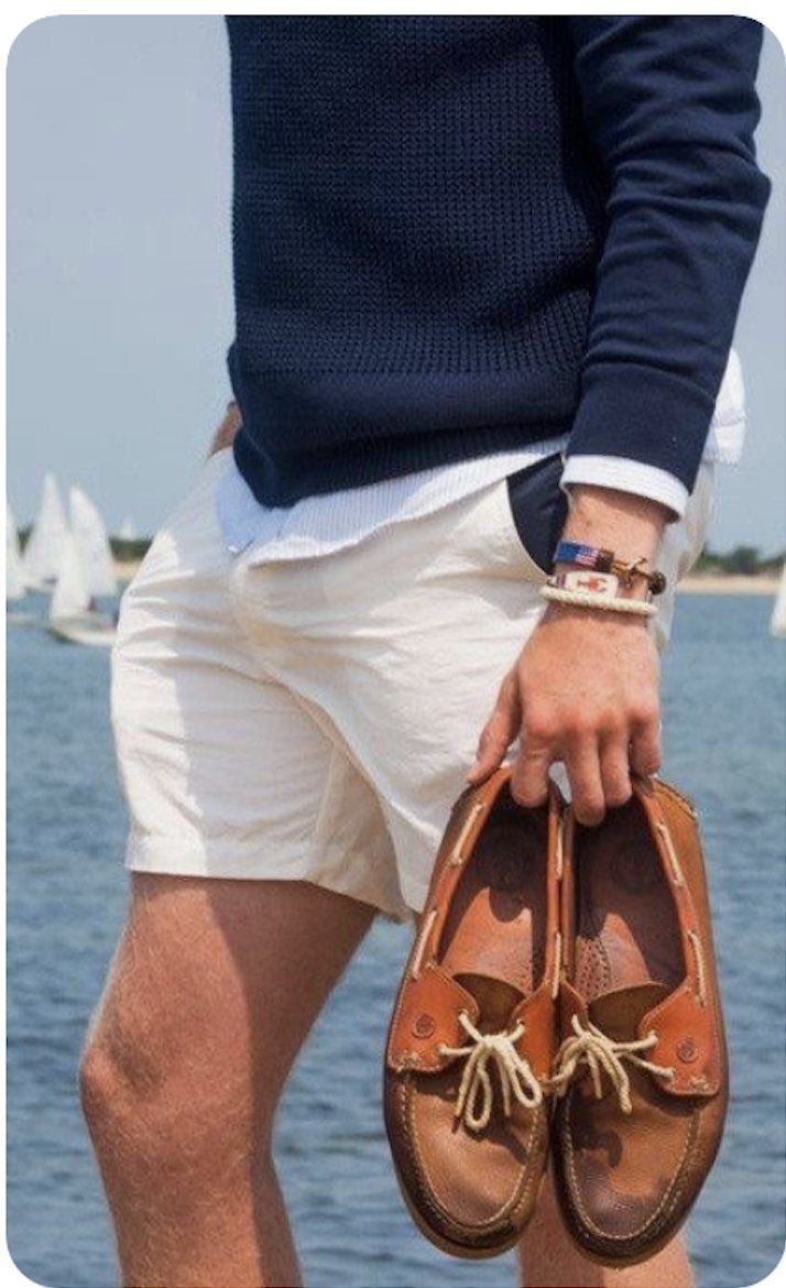 Man wearing a navy sweater, white shirt, and cream shorts holding brown leather boat shoes.