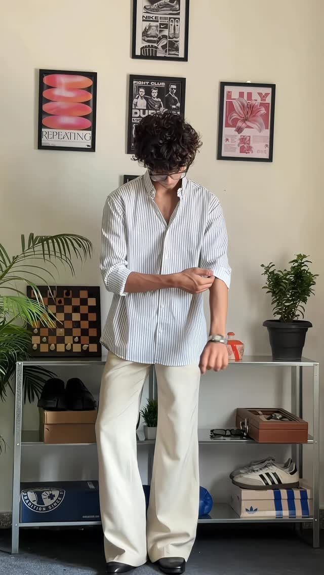 A person wearing a striped button-down shirt and cream wide-leg trousers standing indoors.