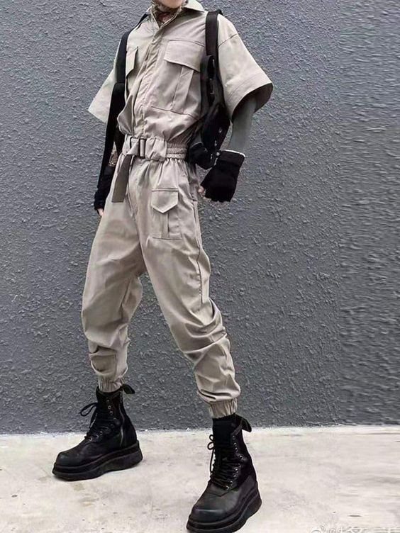 Person wearing a beige utility jumpsuit with black combat boots and a black backpack against a grey wall.