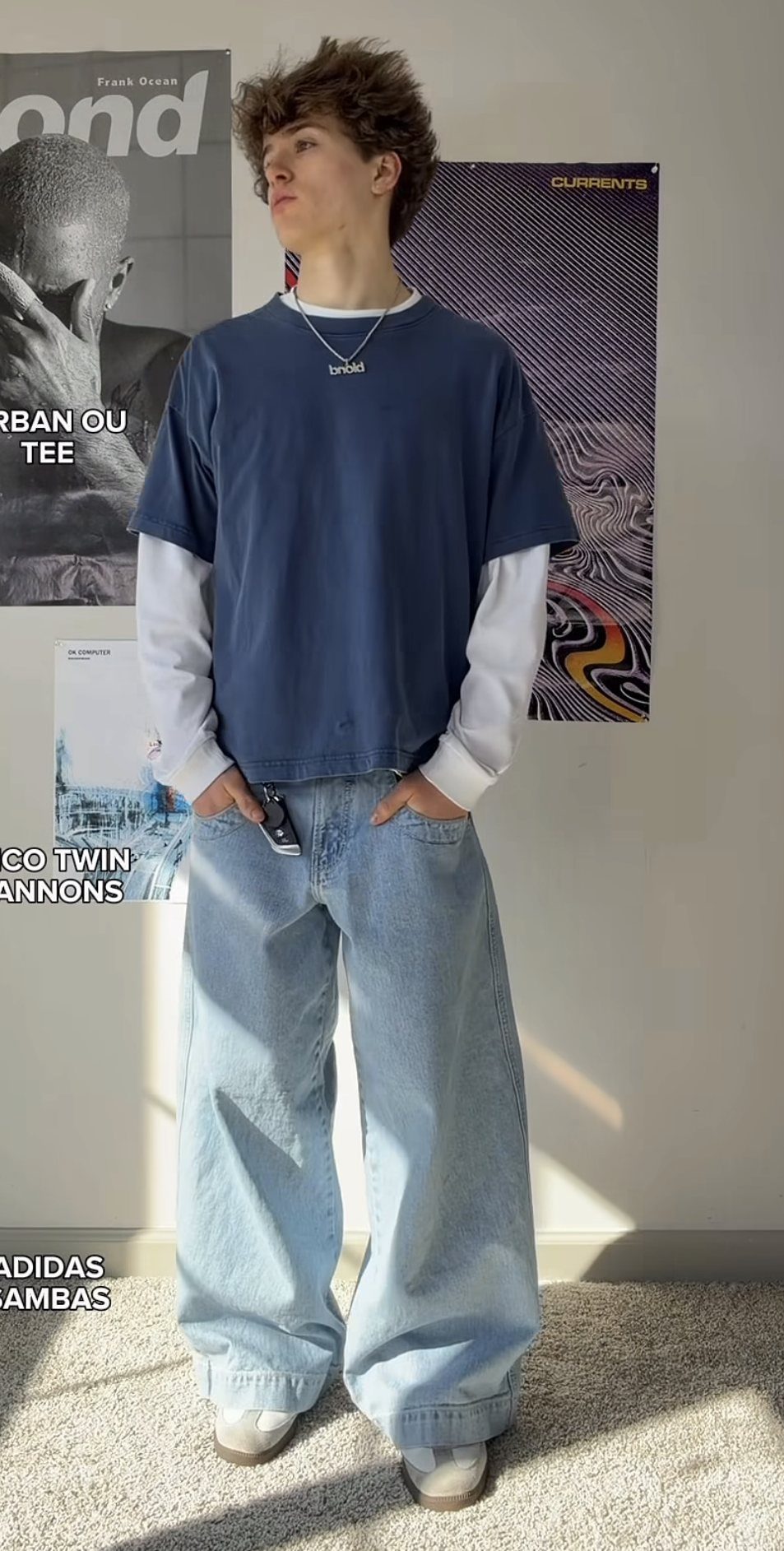 Young man posing in an oversized blue t-shirt, white long-sleeve shirt, and baggy light-wash jeans.
