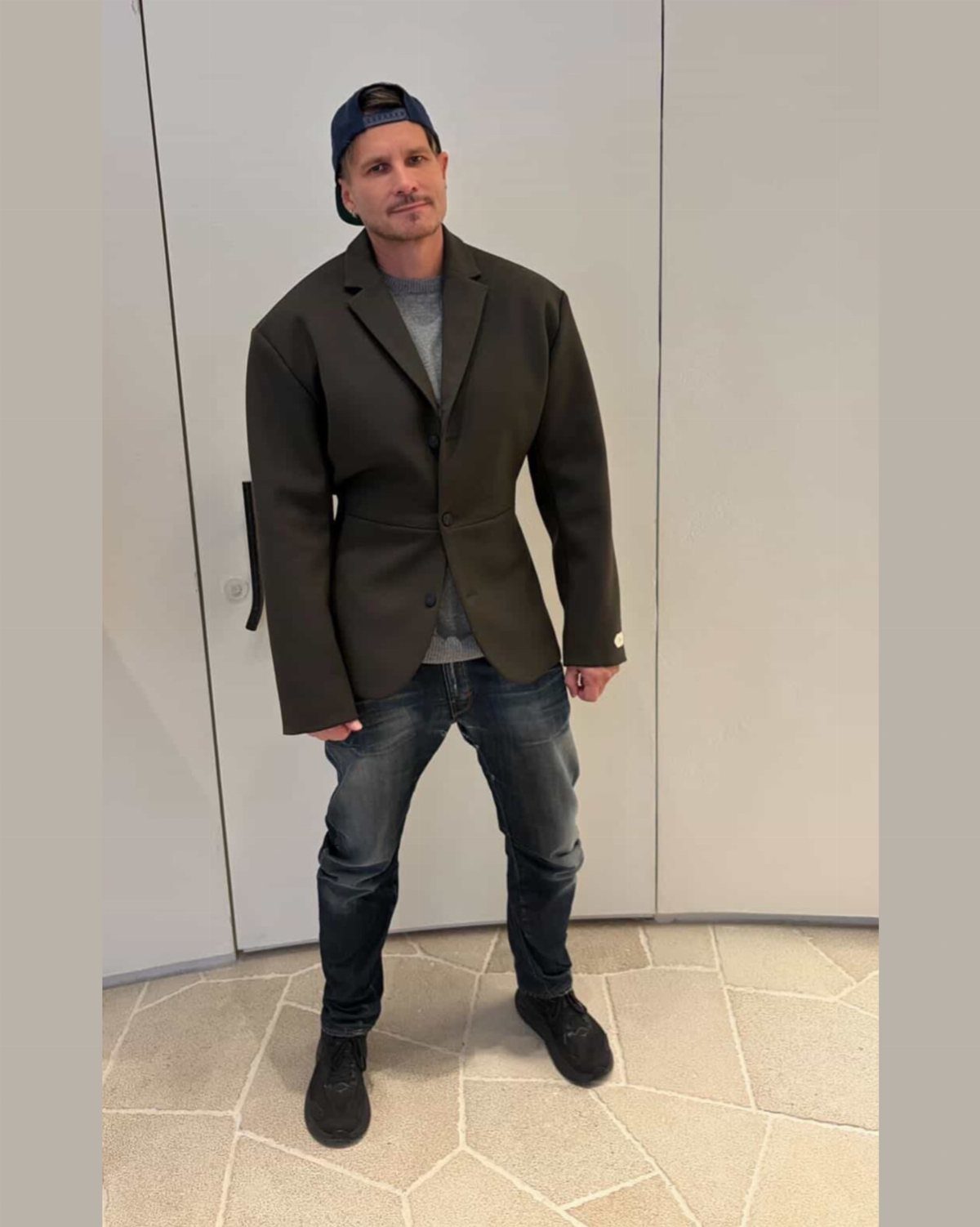 A man posing in a dark olive green statement blazer, grey shirt, and blue jeans.