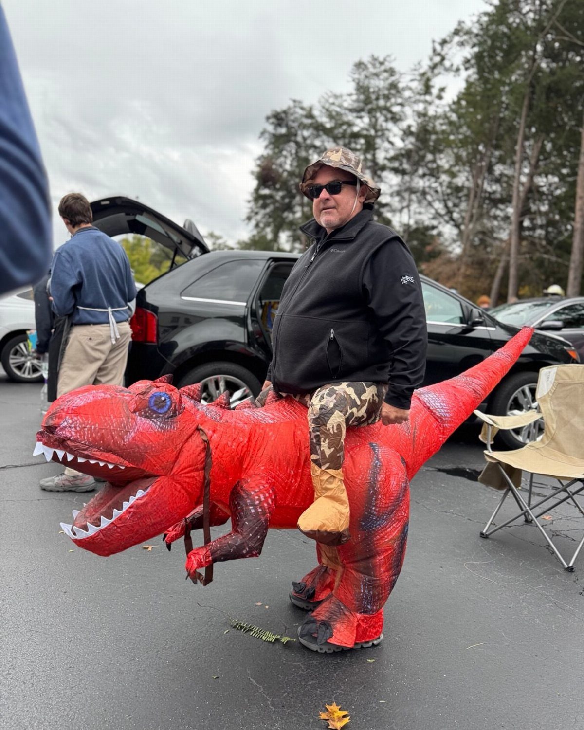 A man riding a large inflatable red T-Rex costume in a parking lot.