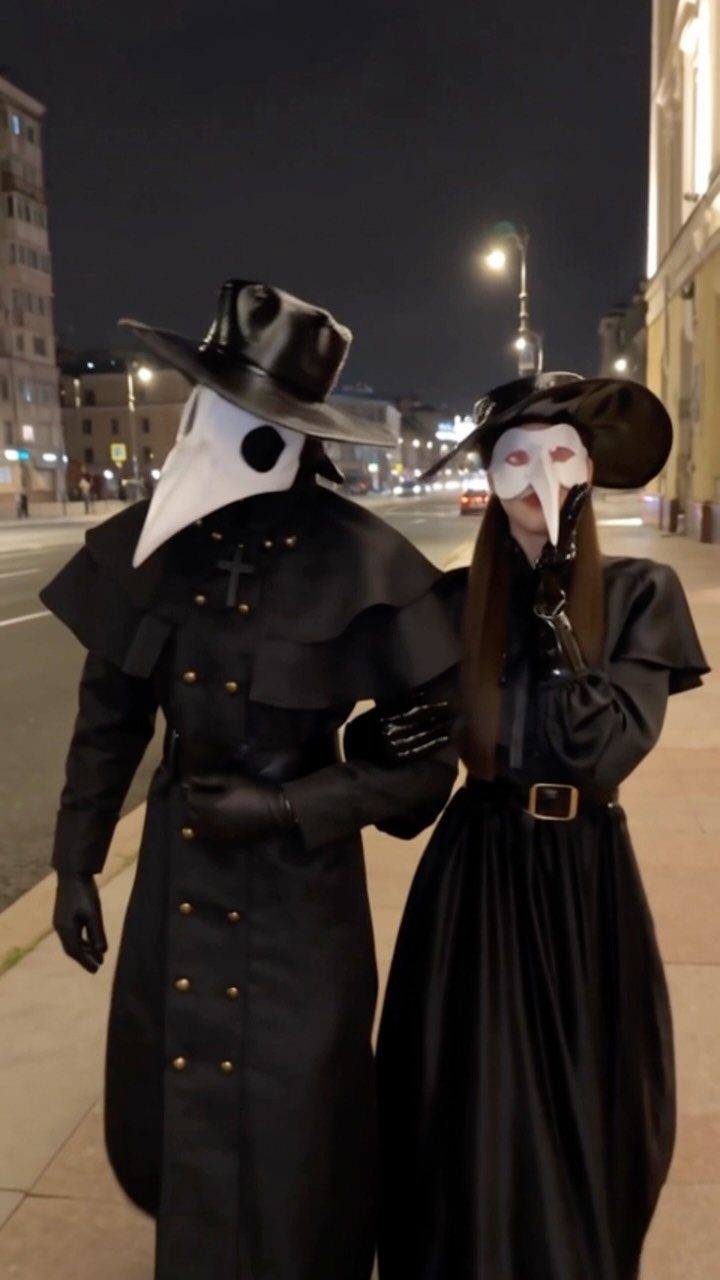 Two people dressed in black plague doctor costumes with bird masks standing on a city street at night.