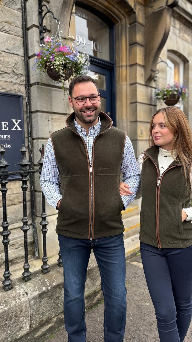 A man and woman smiling while wearing olive green fleece vests outdoors.