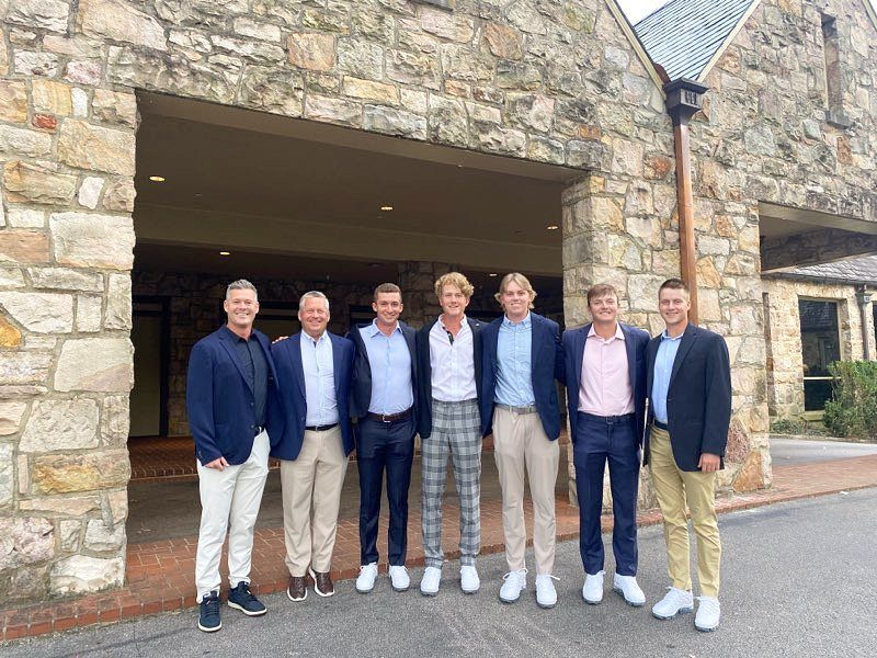 Seven men posing outdoors in blazers, button-down shirts, and trousers in front of a stone building.