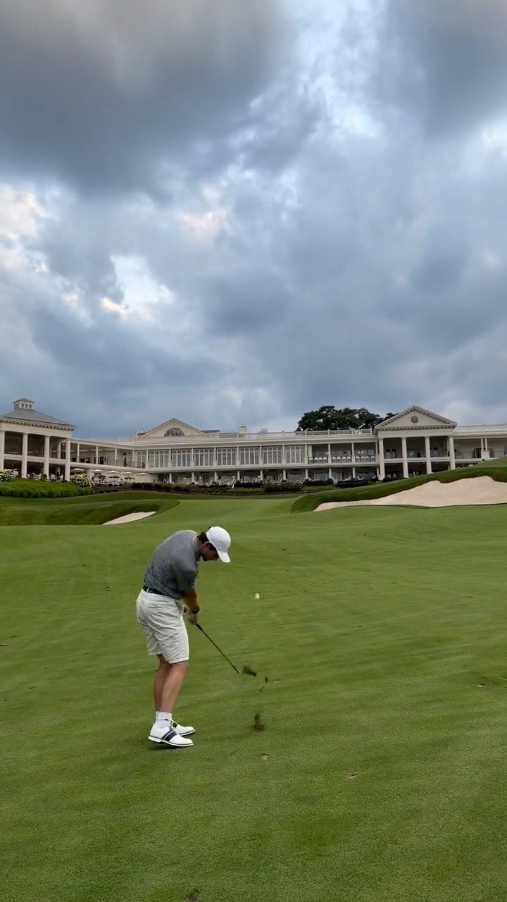 A man in golf attire swings a club on a green course near a clubhouse.