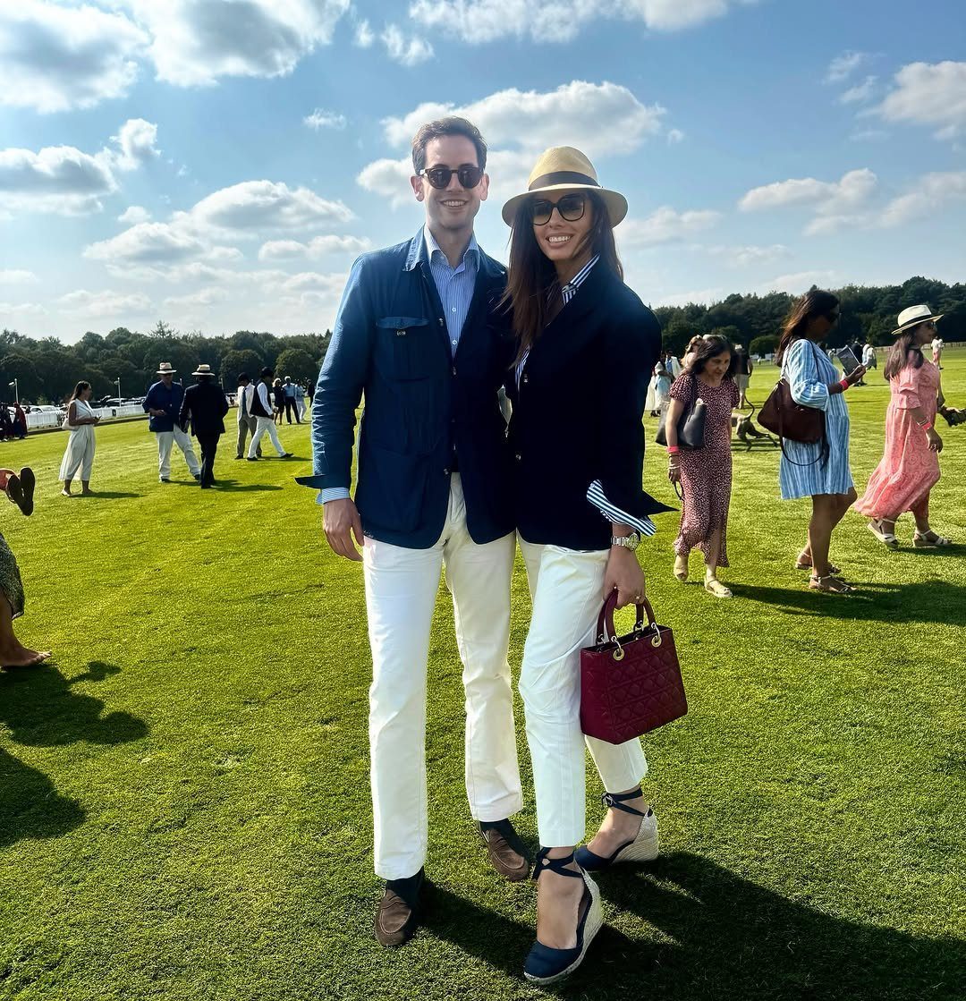 A man and woman in navy blazers and white trousers posing on a green lawn.