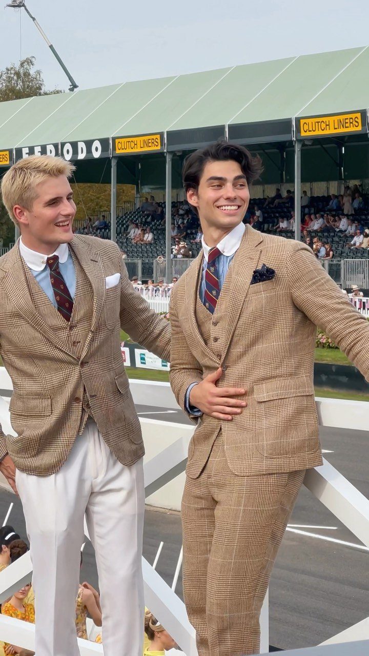 Two young men posing in coordinated tan patterned blazers and preppy attire at an outdoor event.