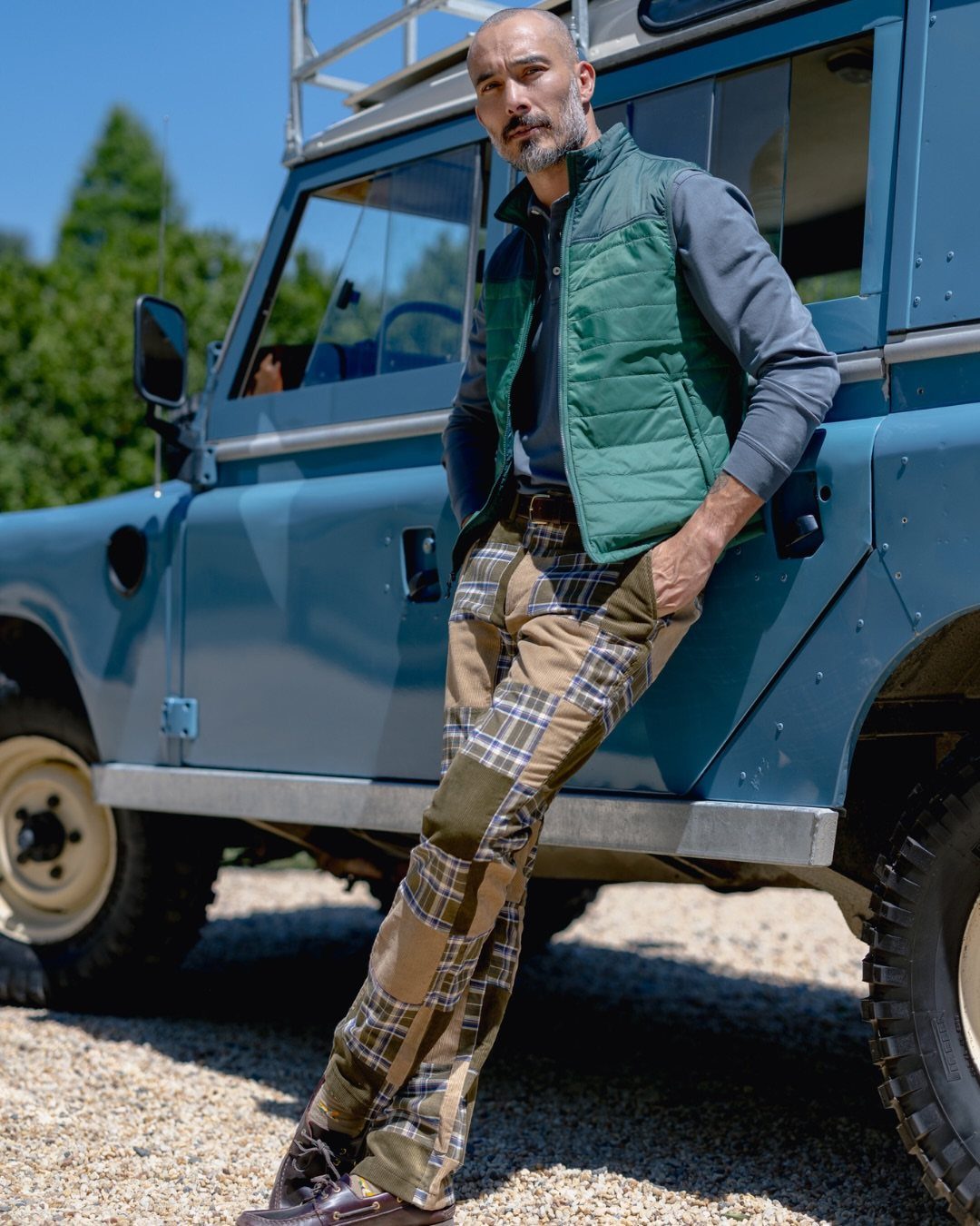 Man in a green vest and plaid trousers leaning against a blue vintage vehicle.