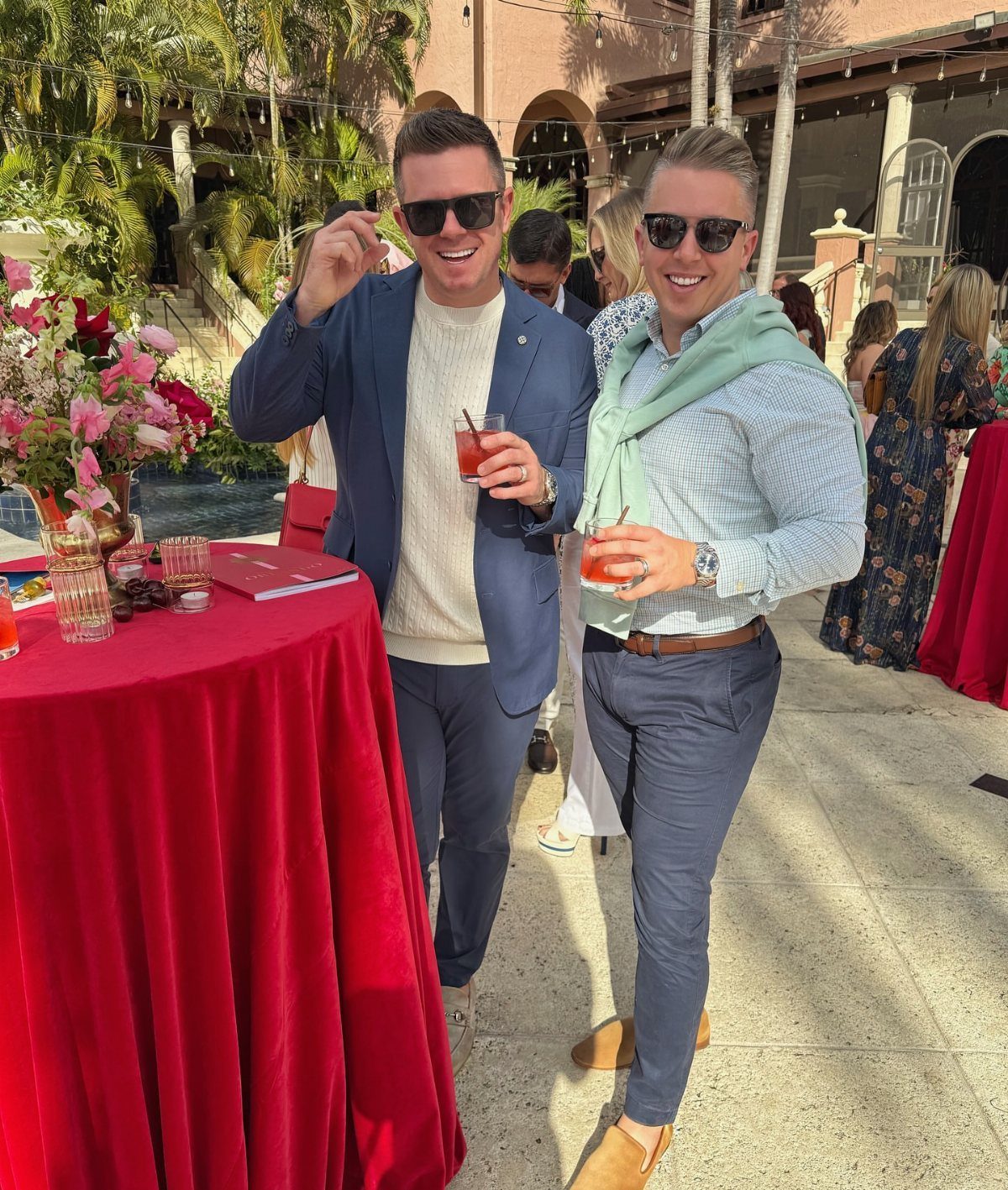 Two men in smart casual attire smiling and holding drinks at an outdoor social event.
