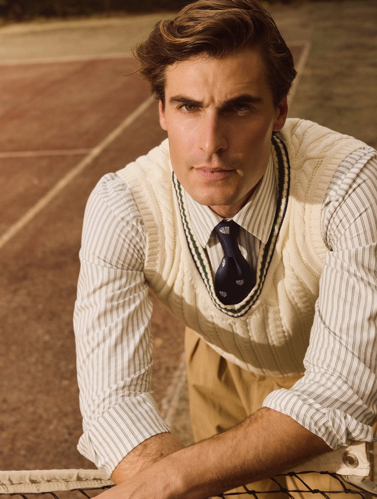 Man in a cable-knit sweater vest and striped shirt leaning on a tennis net.