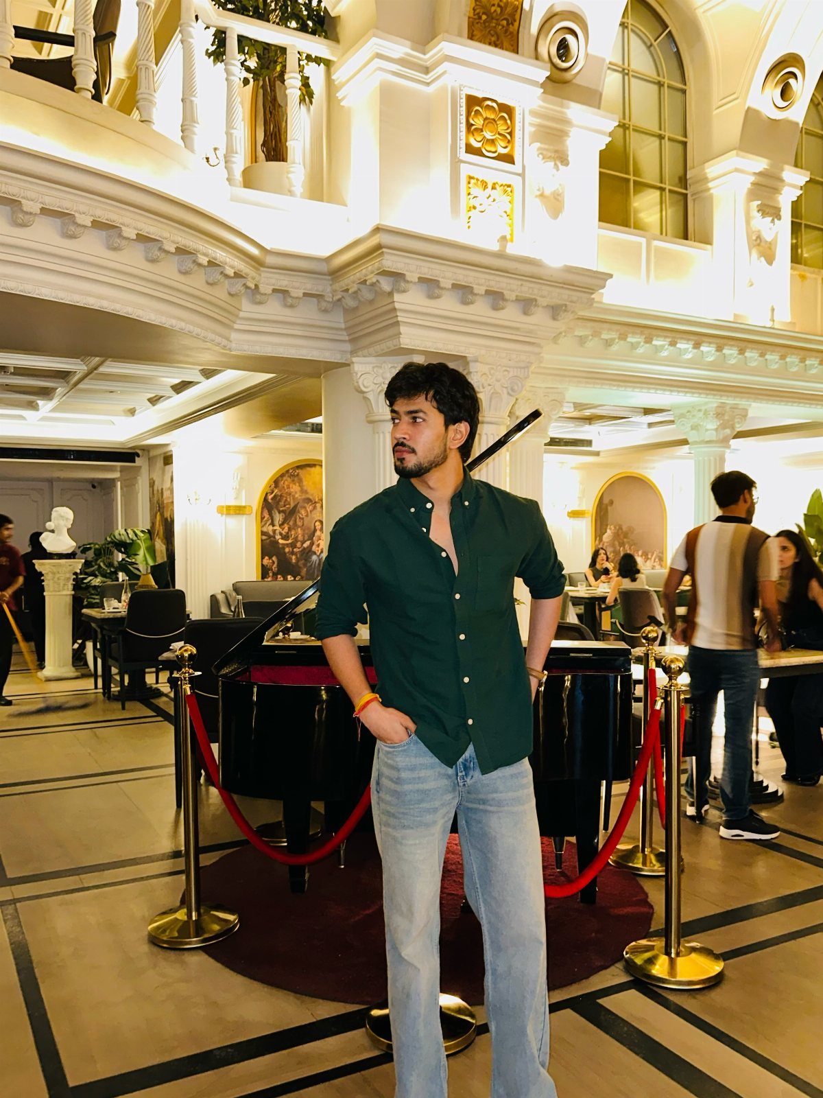 Man wearing a dark green button-up shirt and light blue jeans in a grand hotel lobby.