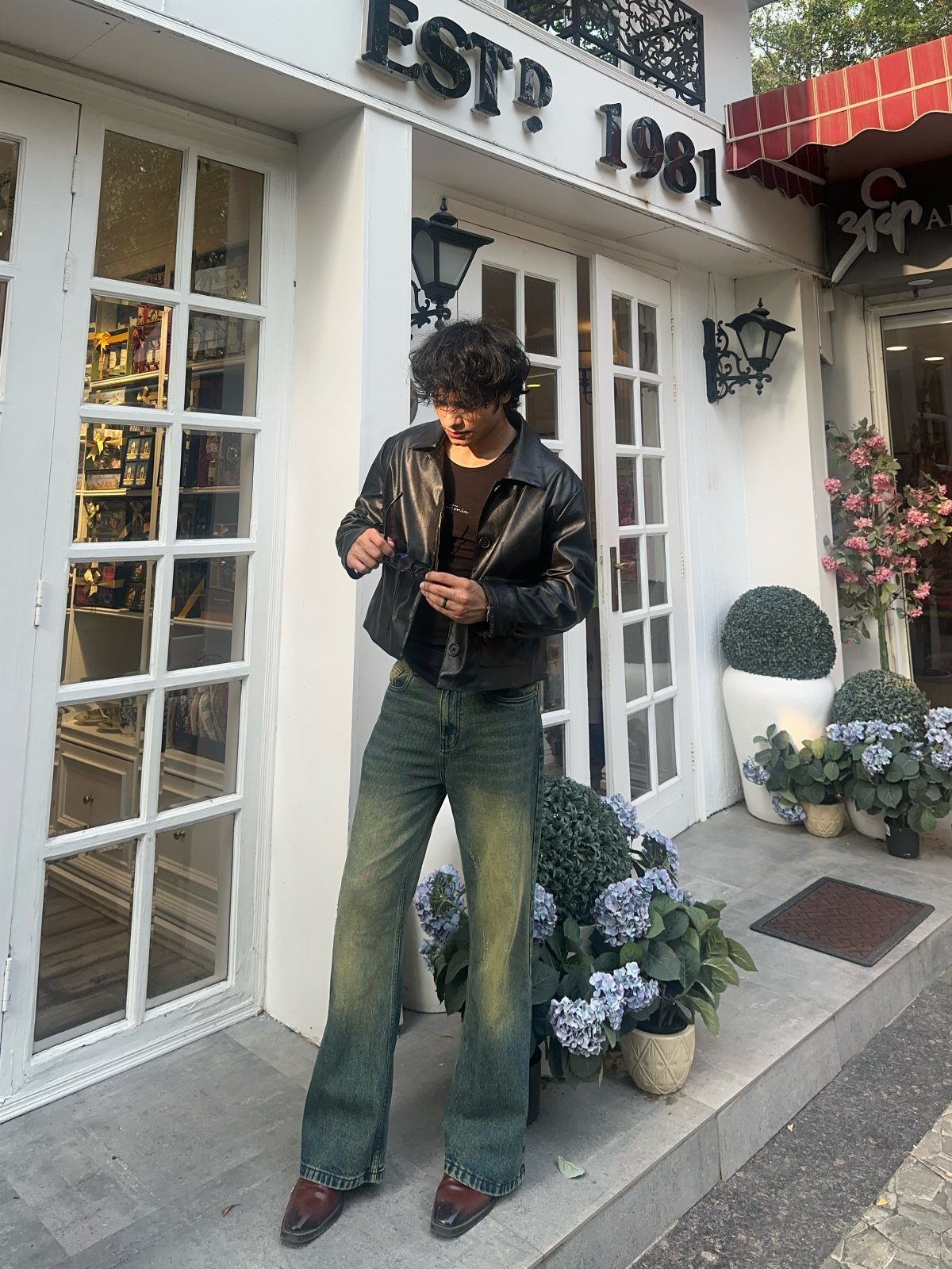 A man wearing a leather jacket and wide-leg denim jeans standing outside a shop.