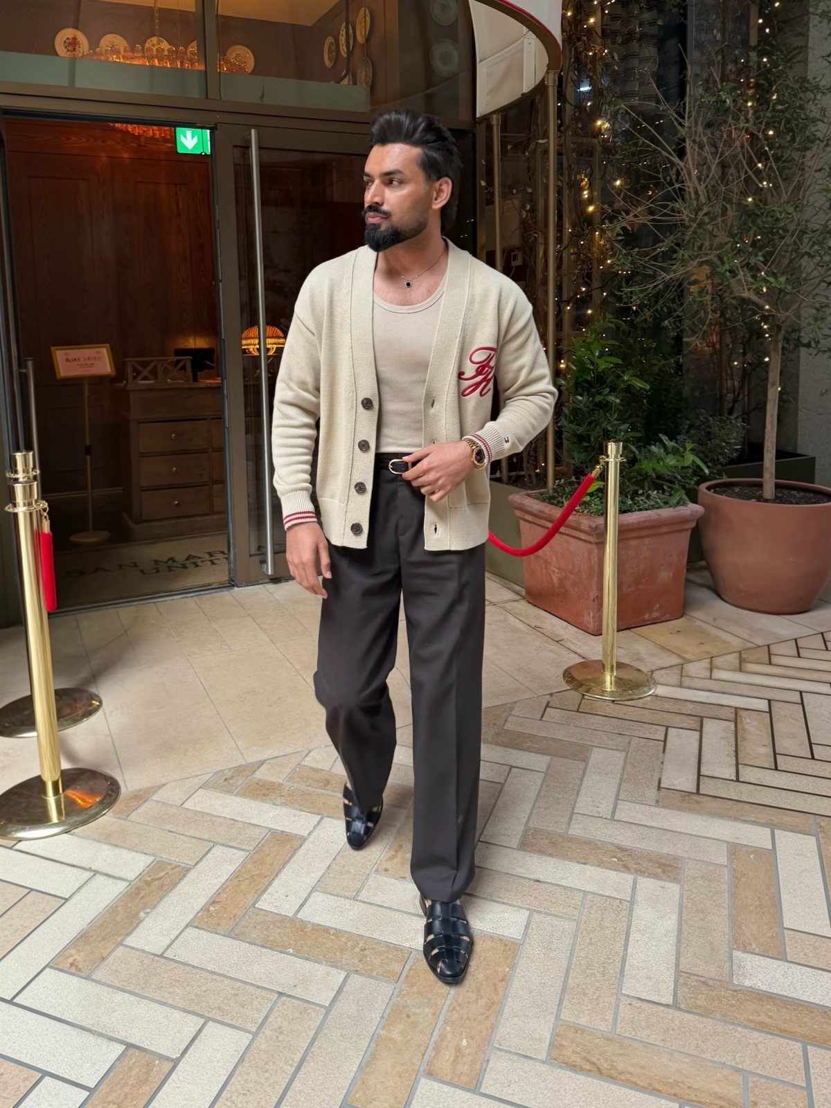 A man wearing a cream knit cardigan and dark trousers standing on a stone walkway.