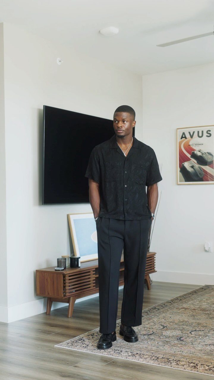 A man posing in a black short-sleeved shirt and black trousers in a living room.