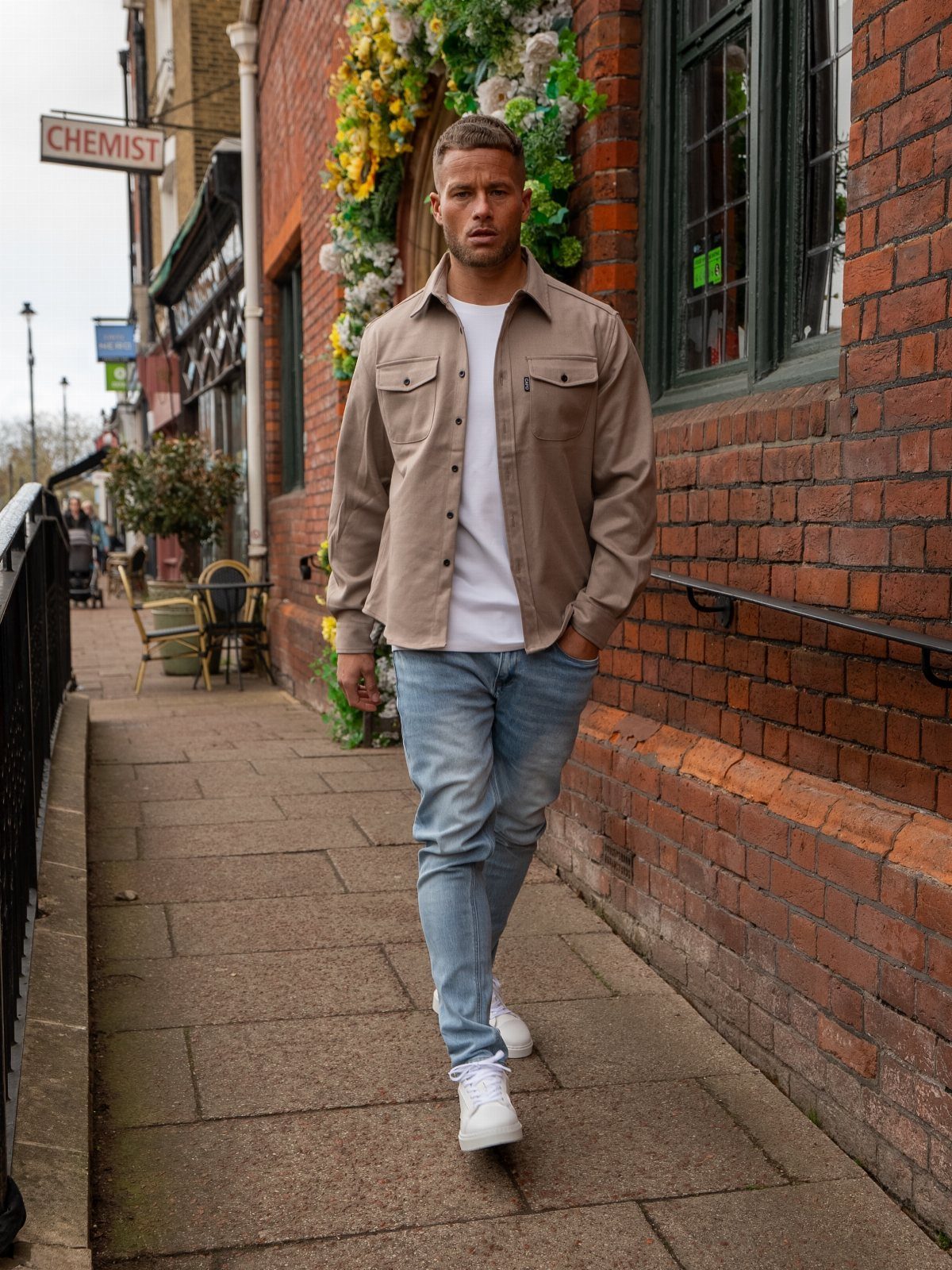A man walking down a sidewalk wearing a tan shirt, white t-shirt, and light blue jeans.