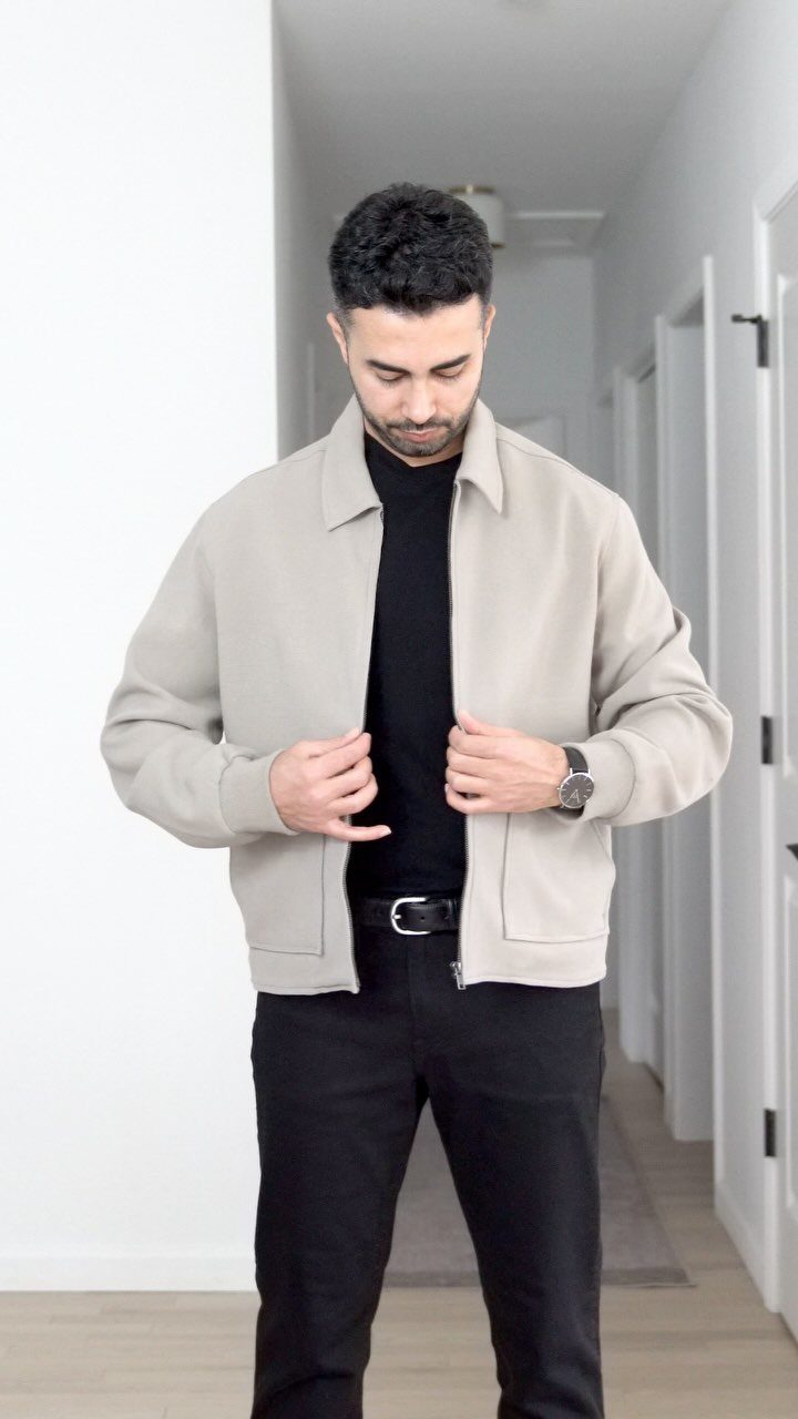 A man wearing a beige jacket, black turtleneck, and black trousers standing in a hallway.
